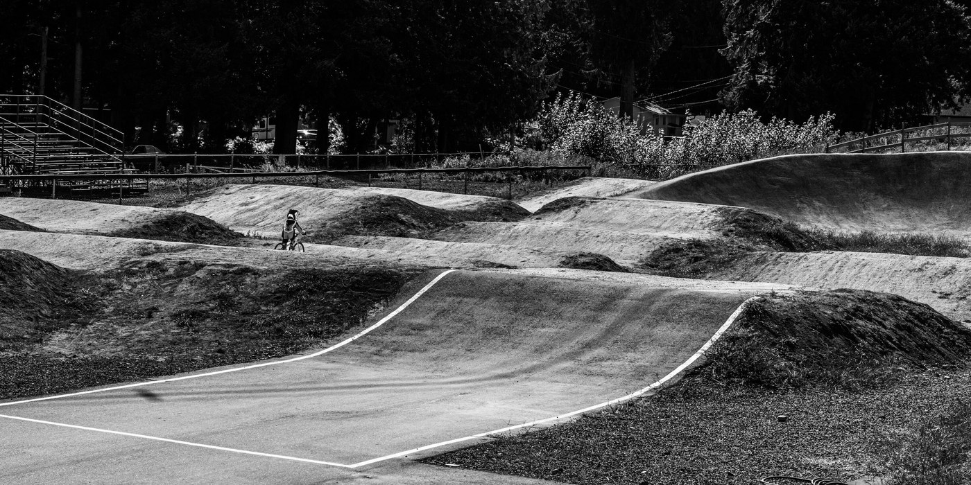 Black and white view across a BMX track, banked dirt berms and a white-edged straight running from the foreground to a distant rider on the far side.