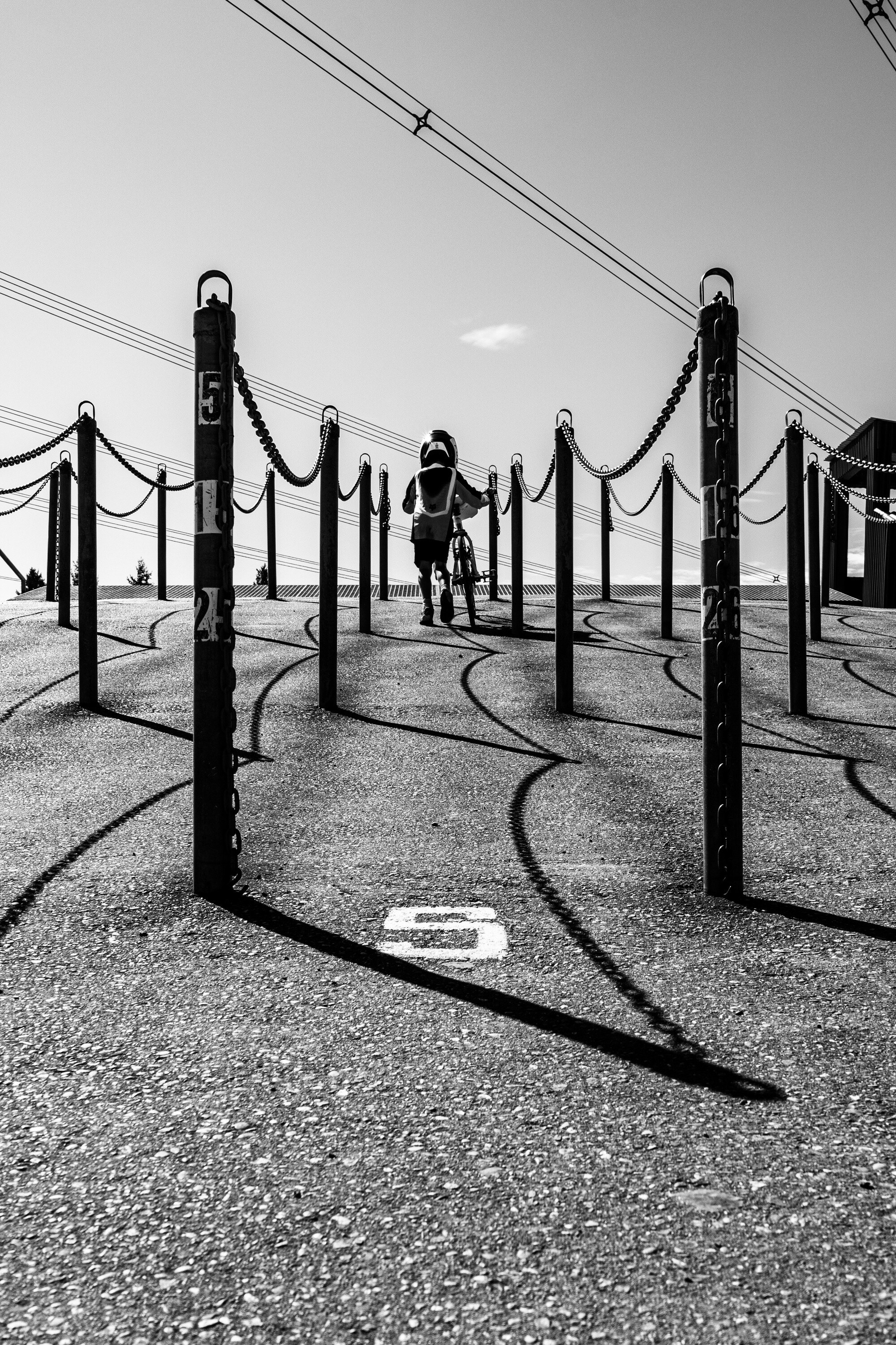 Black and white vertical frame looking up a BMX start hill through two rows of chained posts, tyre lines curving across the asphalt, a single rider at the top.