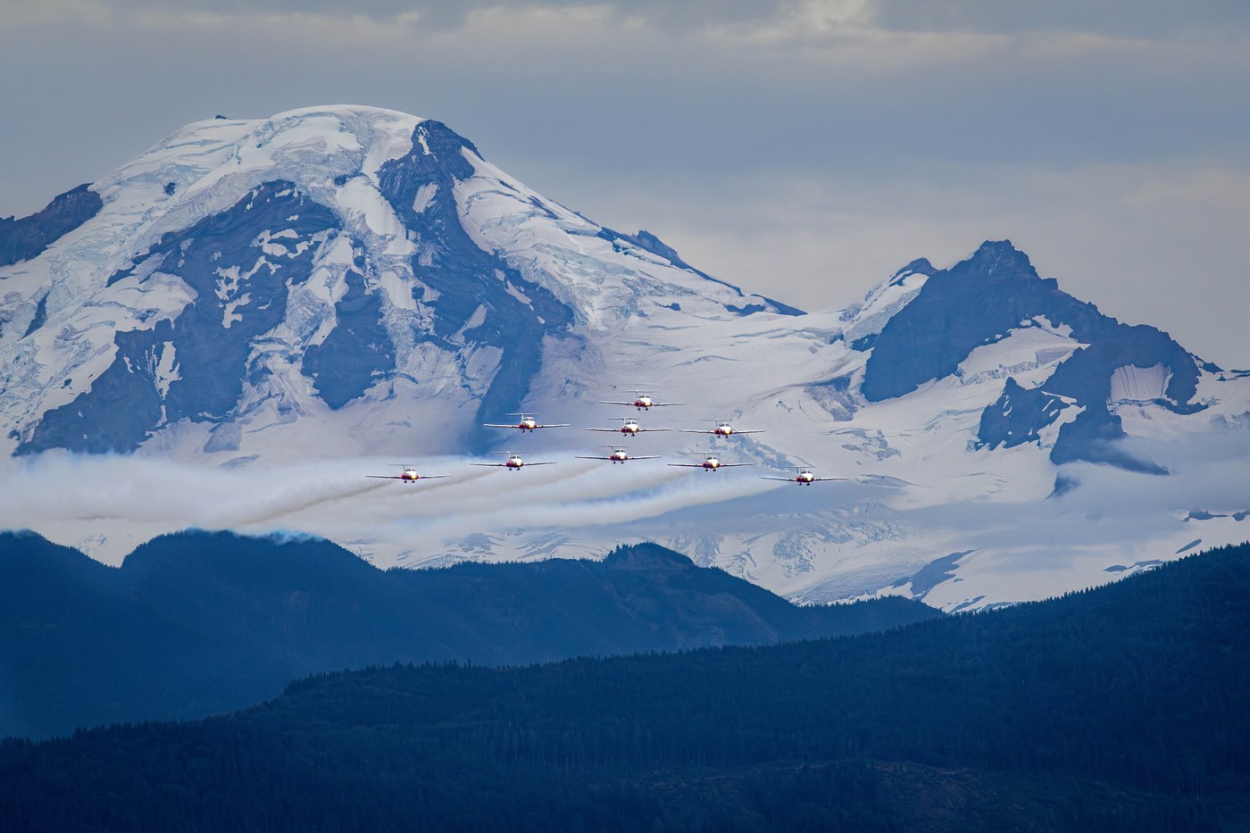 Nine small jets in formation crossing the snow-covered summit ridge of Mount Baker, the mountain filling the frame behind them.