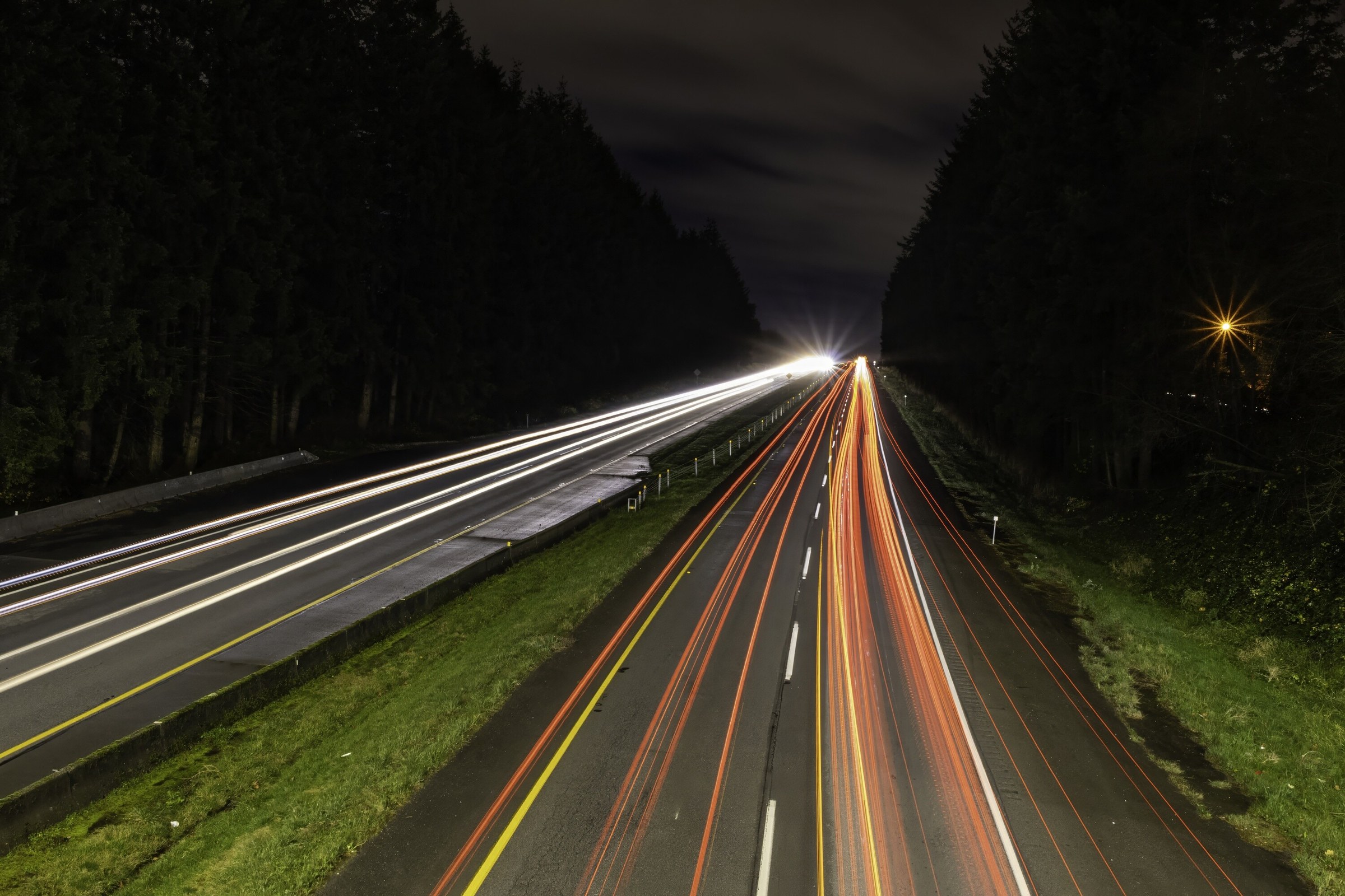A thirty-second exposure down a divided highway at night, white headlight trails on one carriageway and red tail lights on the other, dark forest either side.