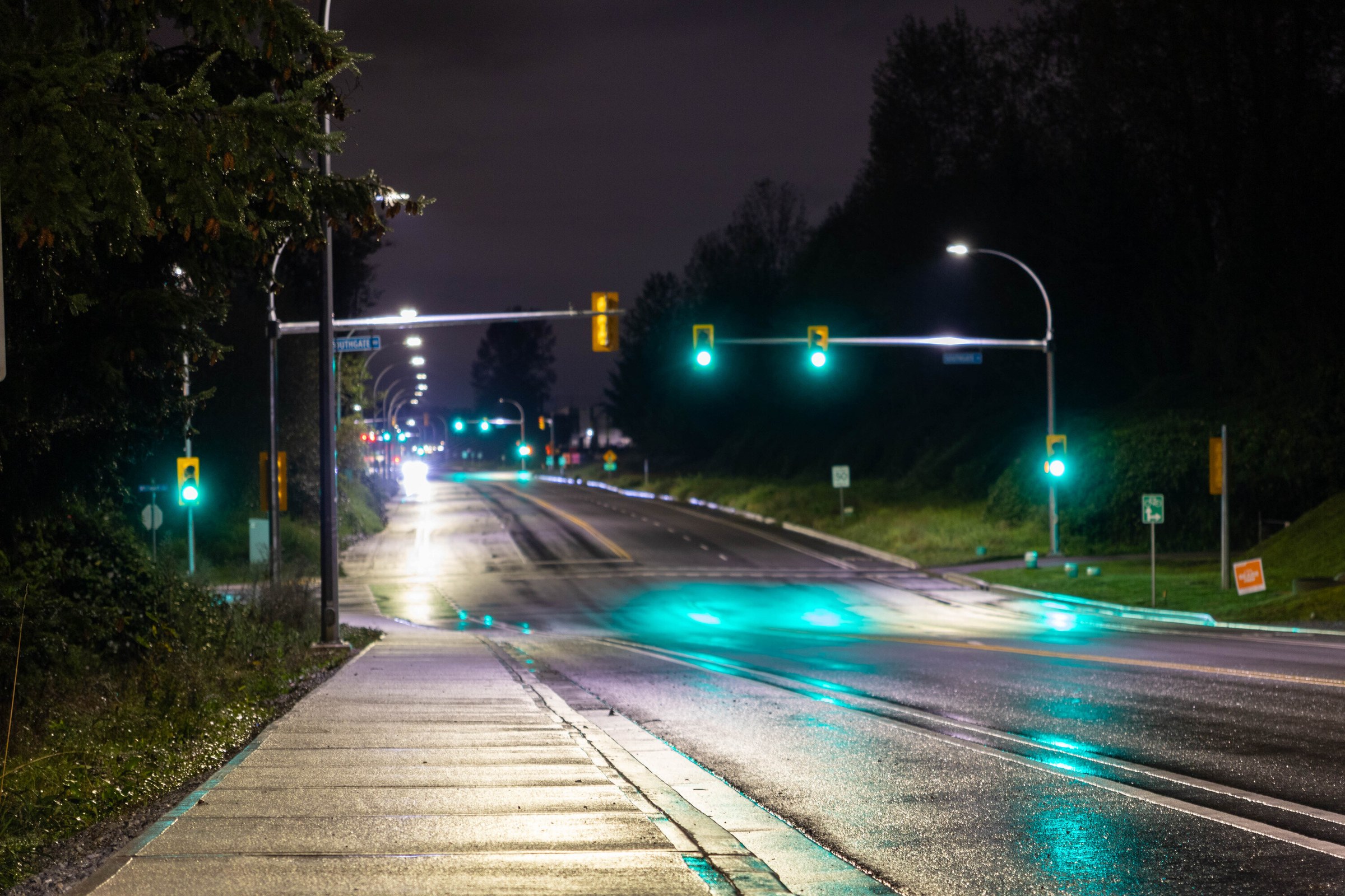 A wet suburban arterial at night, green traffic signals repeated down the road and reflected in long streaks on the asphalt.
