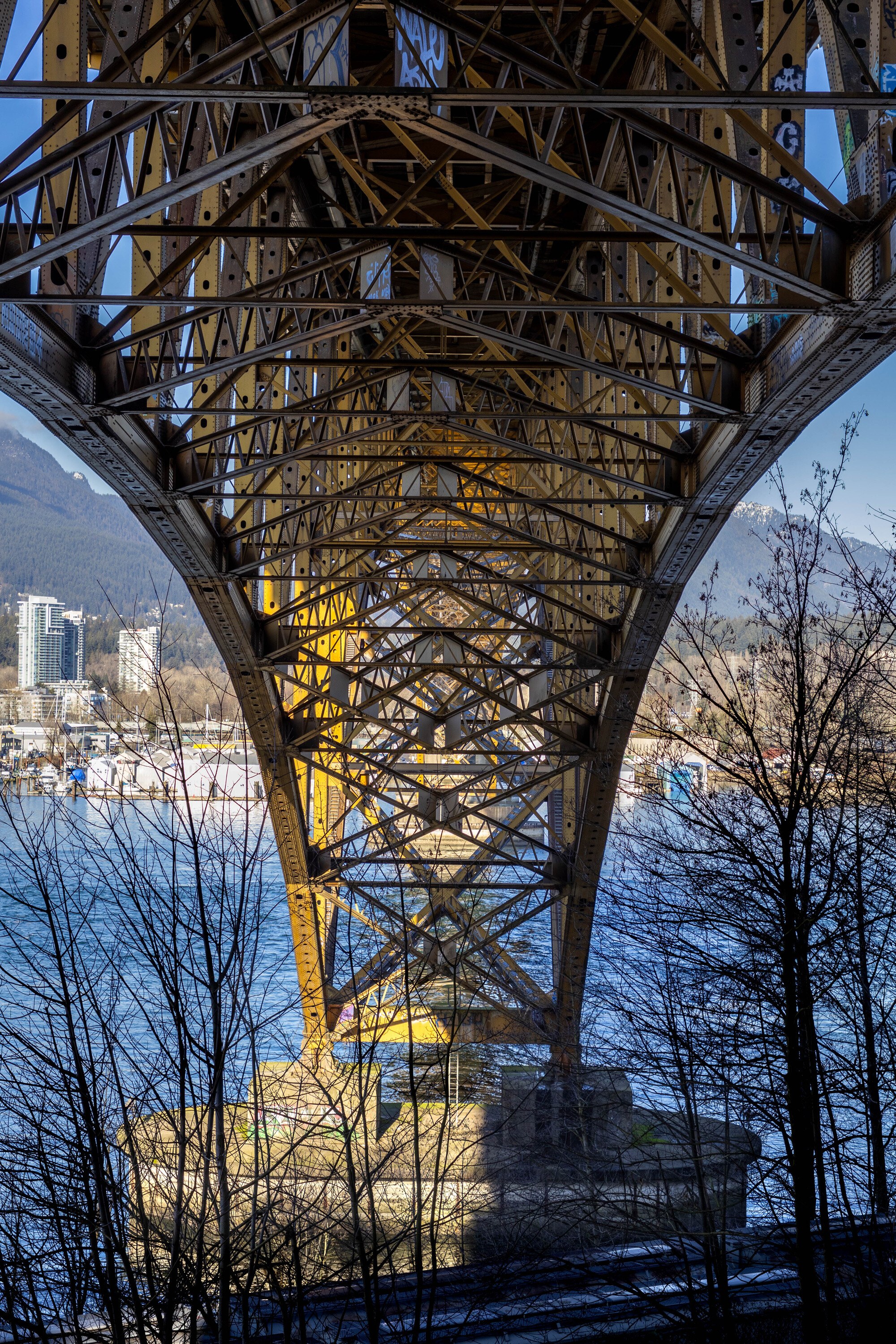 Looking up into the riveted steel underside of the Ironworkers Memorial Bridge, the truss splaying overhead and the inlet visible beneath.