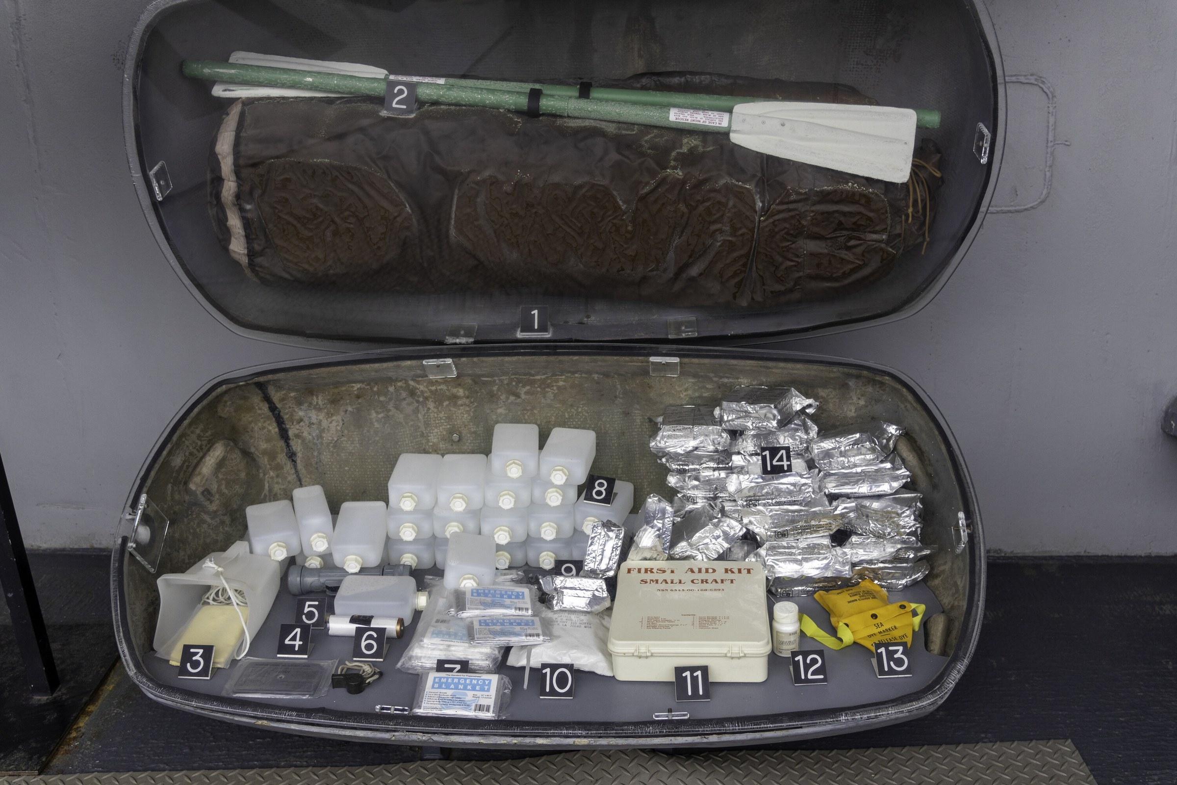 An opened cylindrical life raft container displayed frontally on a museum deck, its contents numbered and laid out in rows.