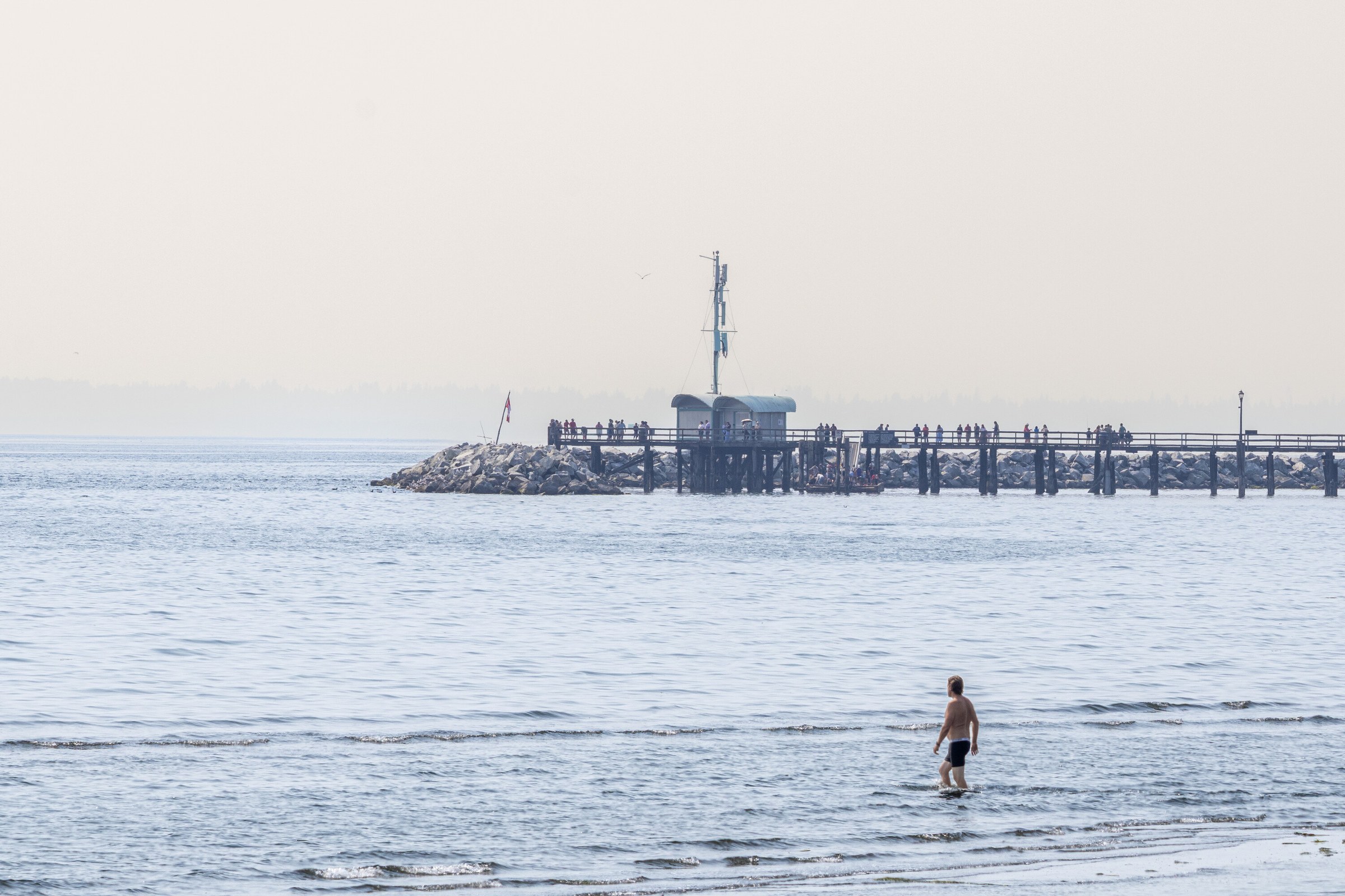 A pale, almost empty frame of White Rock pier and a breakwater across flat water, one small figure standing in the shallows at bottom right.