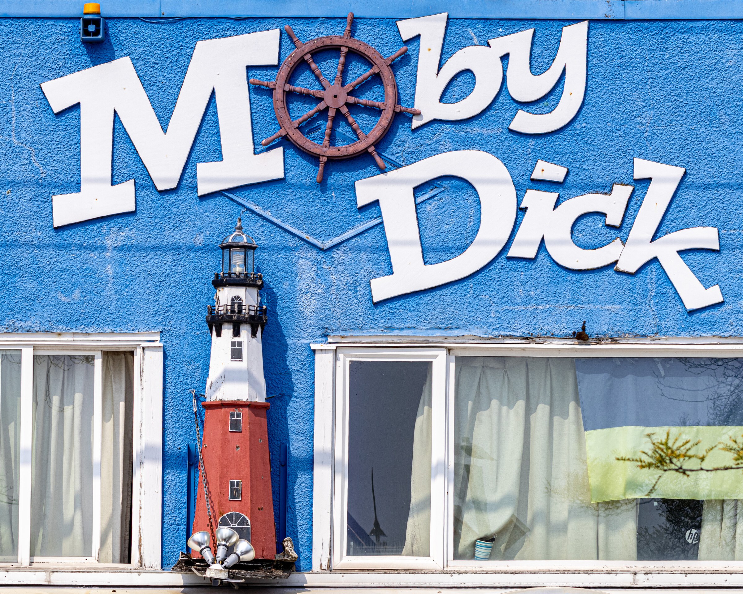 The blue painted front of a fish and chip shop with large white Moby Dick lettering, a model lighthouse between two curtained windows.