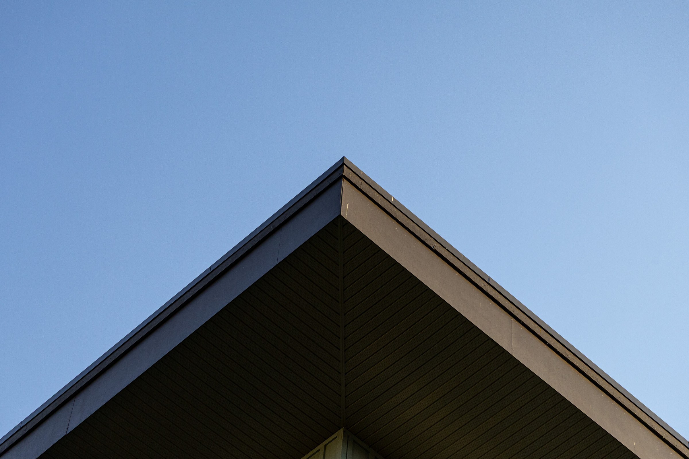 The apex of a roof cutting a hard diagonal V across an otherwise empty pale blue sky.